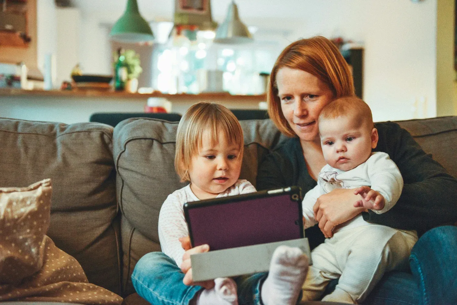 A mother on the couch holding a baby while a toddler looks at a tablet
